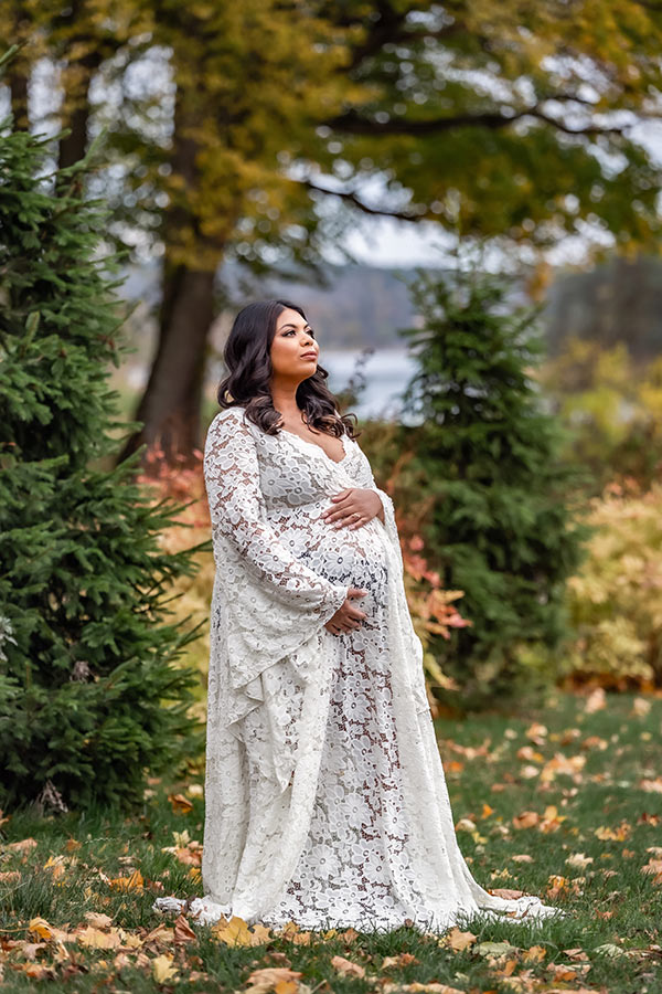 En gravid kvinna står i ett höstlandskap med gula löv på marken. Kvinnan har en vit spetsklänning och håller om magen.