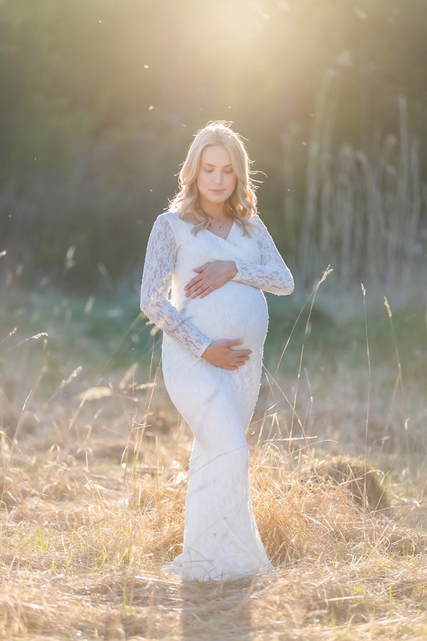 Gravidfotografering där den gravida kvinnan står i motljus på en äng. Kvällssolen glimmar magiskt i de höga grässtråna.