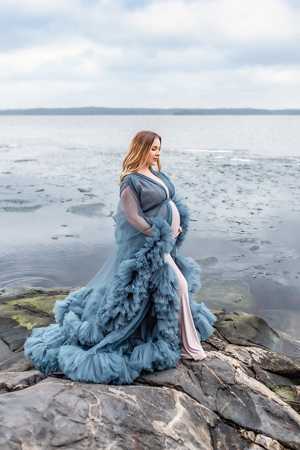En gravid kvinna står på klipporna vid ett isigt hav. Hon tittar stillsamt ner på sin fina mage.