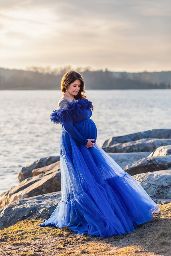 Gravidfotografering en vindig dag vid havet. Kvinnan har en blå klänning.