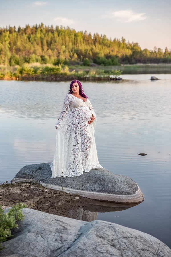 En gravid kvinna står på en stor sten i vattnet. Hon har en vit gravidklänning och lila hår. Det är sommar och kvällsljuset färger världen mjukt.