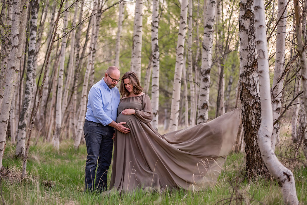 Gravidfotografering i en dunge med björkar. Den gravida kvinnan har en lång klänning som flyger i vinden och hon och den blivande pappan håller om magen.