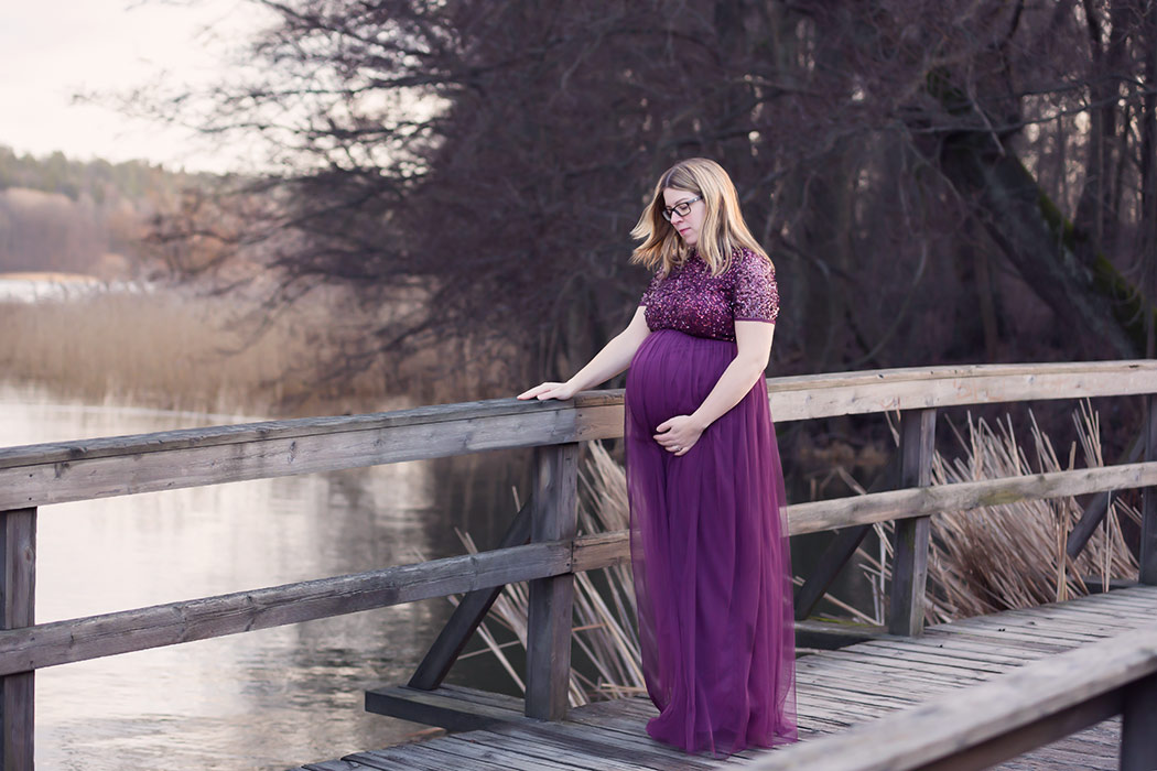En gravid kvinna i en glittrande lila gravidklänning står på en träbro vid vattnet.