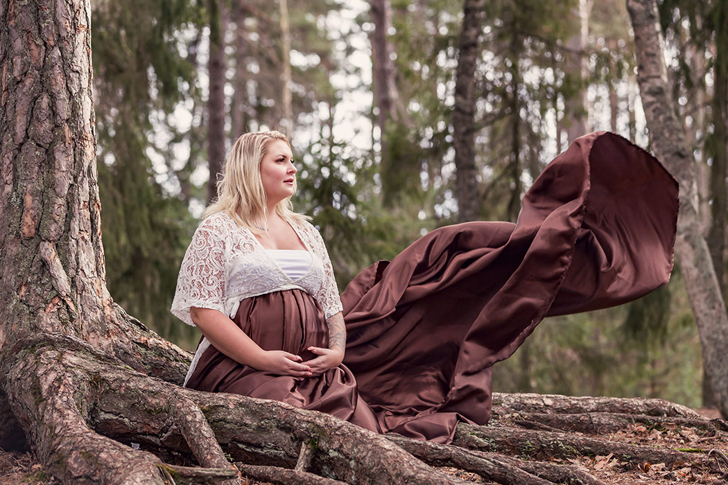 Gravidfotografering i en barrskog. Den gravida kvinnan sitter bland stora rött och hennes bruna satinkjol flyger i vinden.