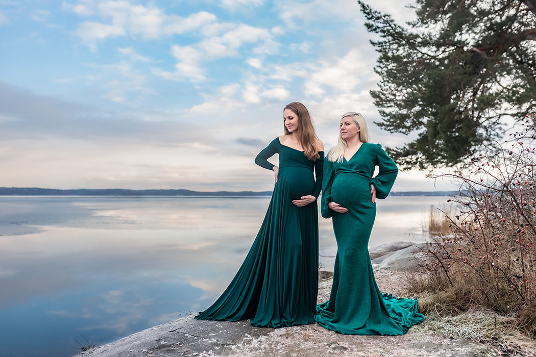 Två gravida systrar i gröna gravidklänningar står på klippor vid havet en kall vinterdag.