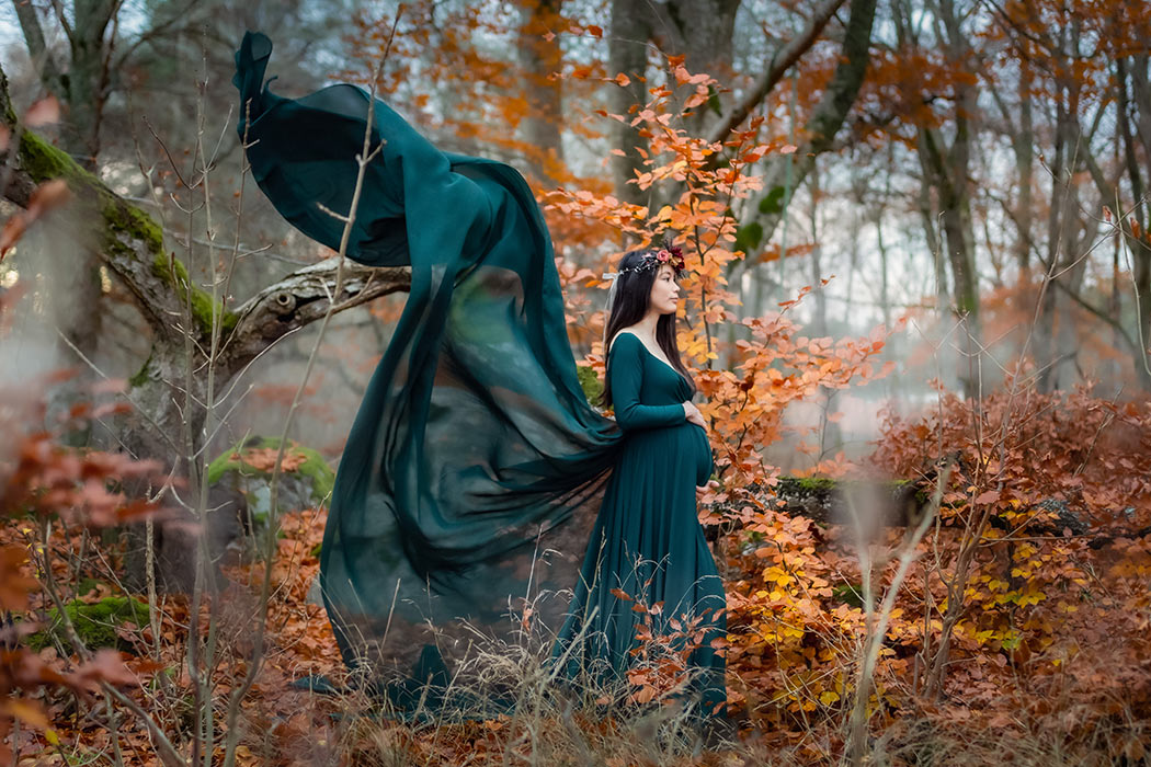 Gravidfotografering i en höstskog där alla löven är vackert orange. Kvinnan har på sig en grön klänning som fladdrar i vinden.