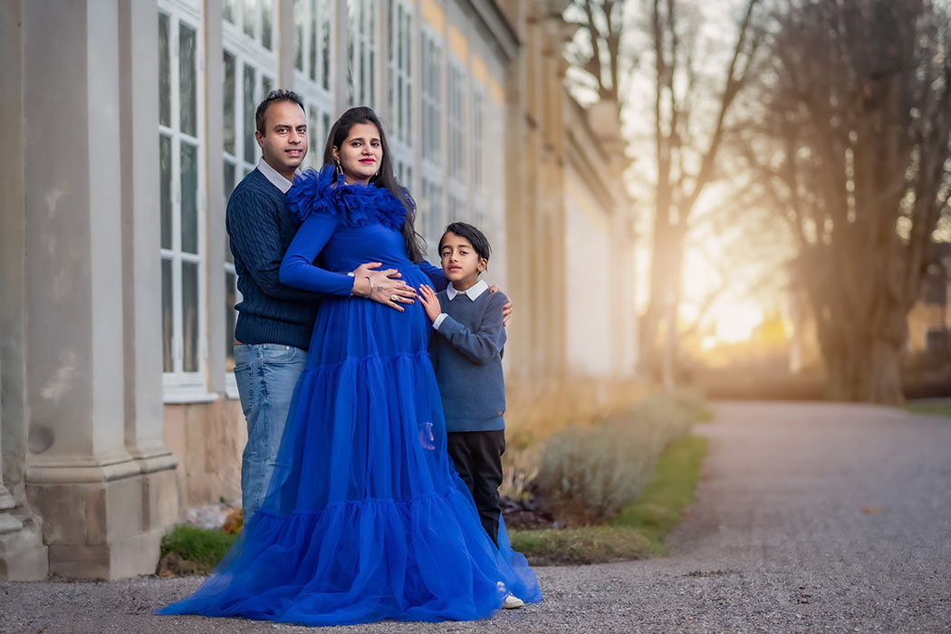 Gravidfotografering i november vid Ulriksdals slott. Den gravida kvinnan har en blå gravidklänning och står tillsammans med sin son och man vid orangeriet. I bakgrunden syns träd utan blad i kvällsljuset.