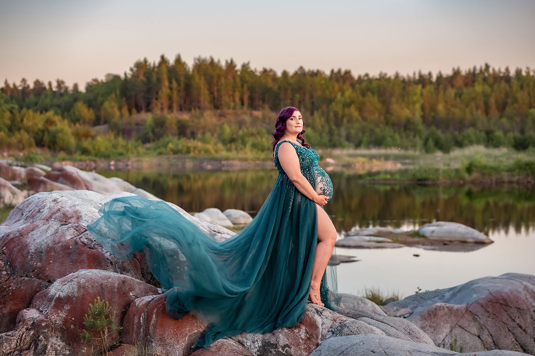 En gravid kvinna står bland vackra klippor framför ett spegelblankt vatten. Hennes gröna glittrande gravidklänning flyger i vinden.