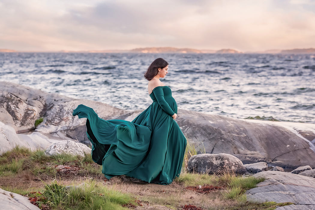 En gravid kvinna i grön klänning står bland klipporna vid havet. Himlen är färgad av kvällsljuset. Den gröna klänningen flyger i vinden.