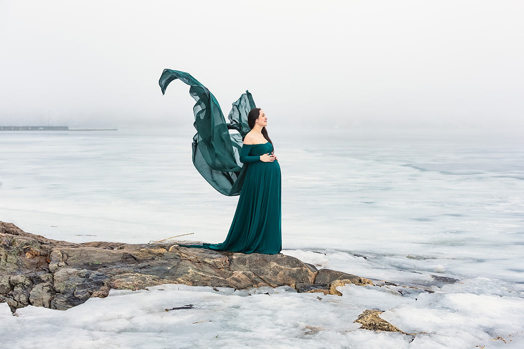 Gravidfotografering en vinterdag när dimman ligger tät. Kvinnan har på sig en grön klänning med släp som flyger som fjärilsvingar.