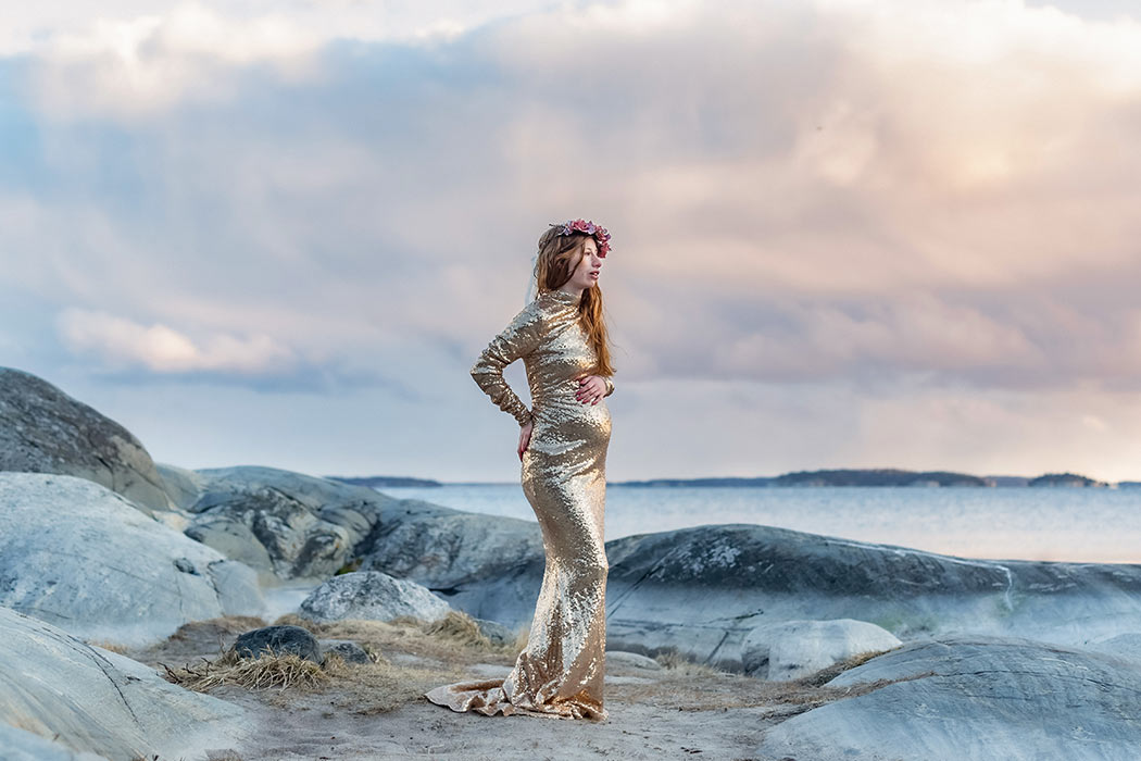 Gravidfotografering med dramatisk himmel bland klipporna vid havet. Kvinnan har en guldklänning och rosa blomsterkrans.