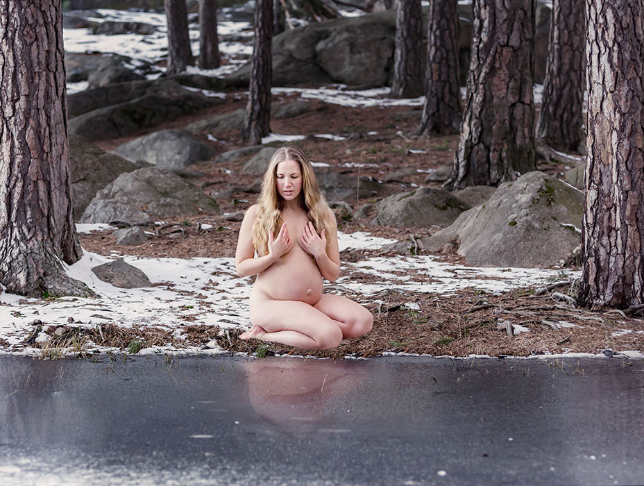 En naken gravid kvinna sitter vid en istäckt sjö. I bakgrunden syns stora stenar och trädstammar.