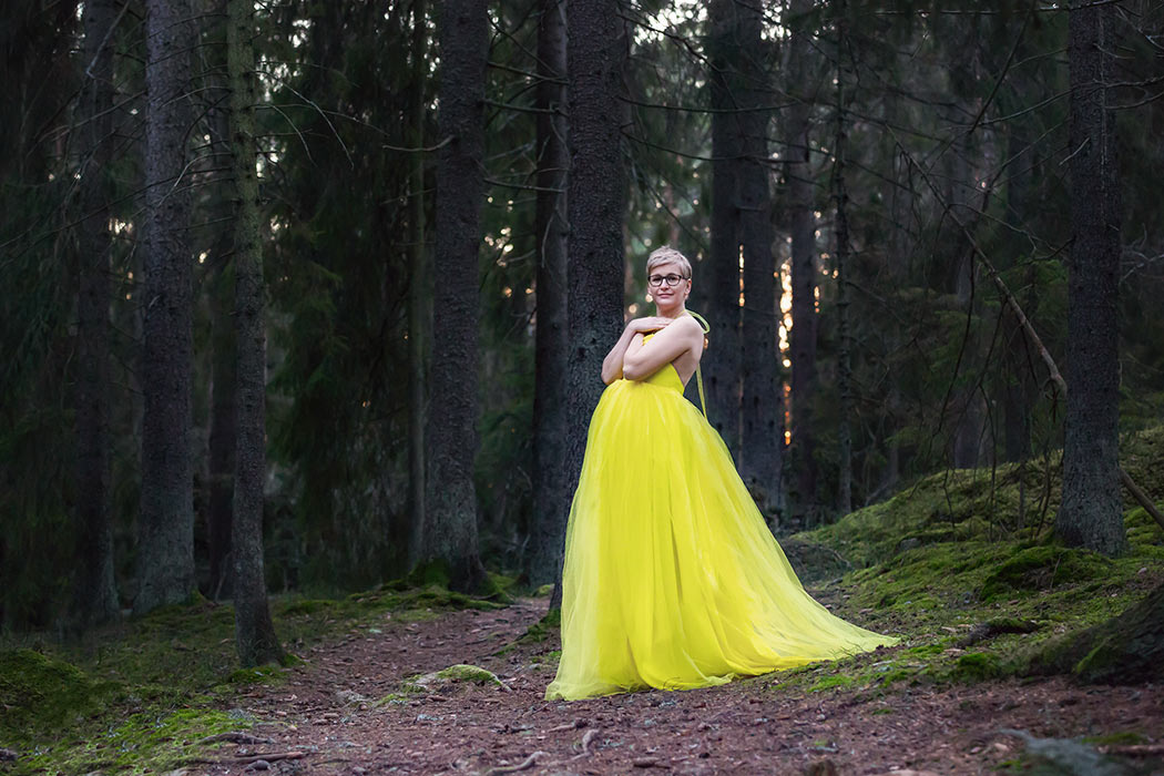 En vacker gravid kvinna i gul klänning står på en stig i en barrskog.