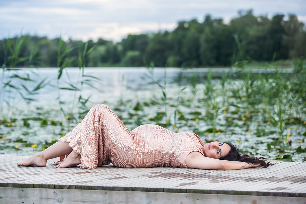 Det är sommar och den gravida kvinnan ligger på en brygga och tittar in i kameran. I bakgrunden syns näckrosor på sjön och gröna träd på andra sidan vattnet.