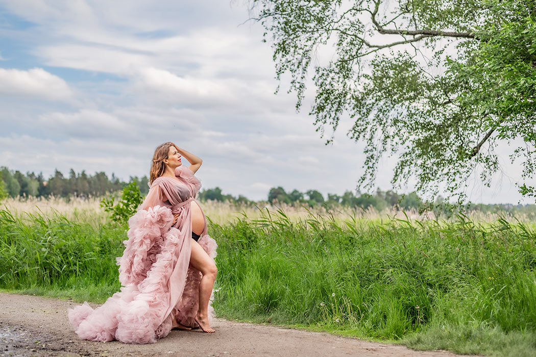 En gravid kvinna går på en grusgång bredvid en äng med höga gräs. På sig har hon en rosa tyllklänning där magen visas.