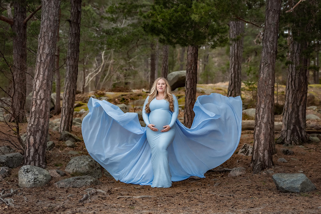 En gravid kvinna står i en barrskog i en blå gravidklänning med långt släp som flyger i luften.