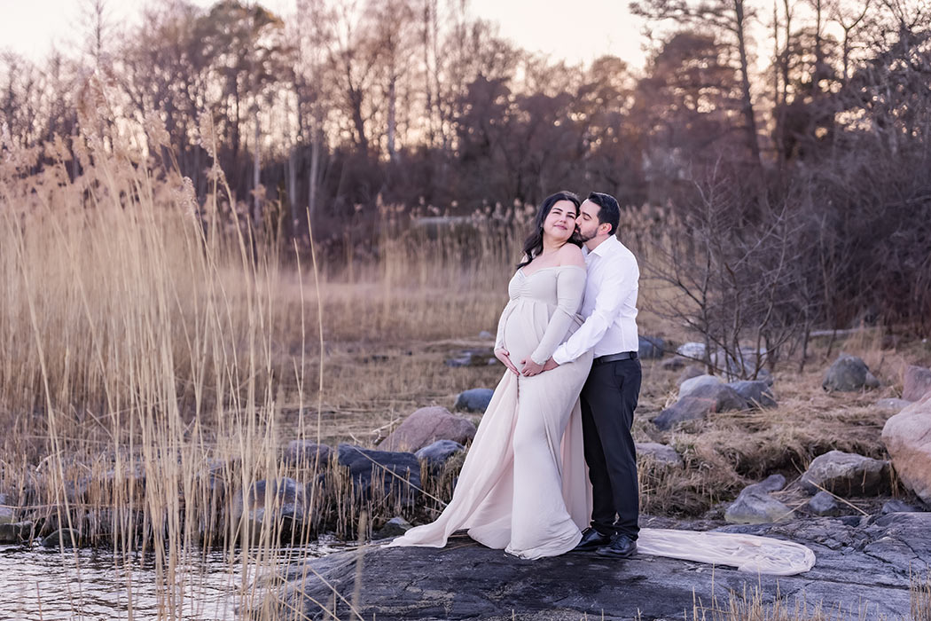 Gravidfotografering i kvällsljuset bland vass vid havet. Paret håller om varandra och mannen pussar kvinnan på kinden.