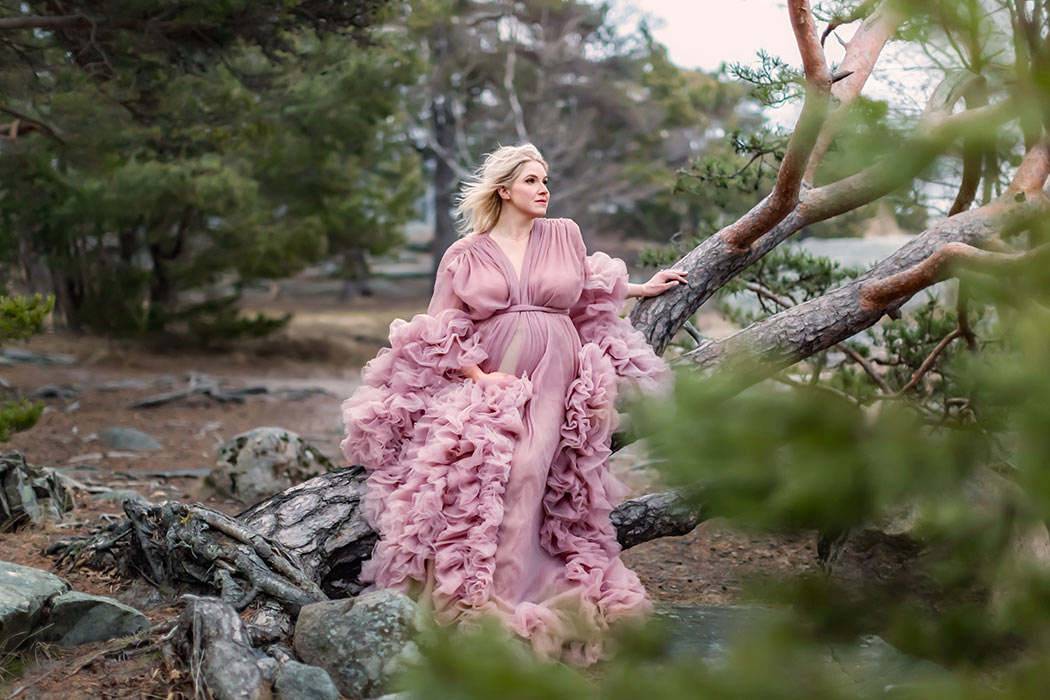 Gravidfotografering med en vacker gravid kvinna som har på sig en rosa tyllklänning och sitter på ett liggande träd.