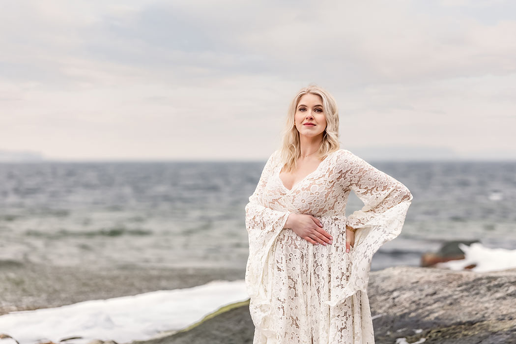 En vacker gravid kvinna i vit spetsklänning står vid havet och tittar in i kameran.