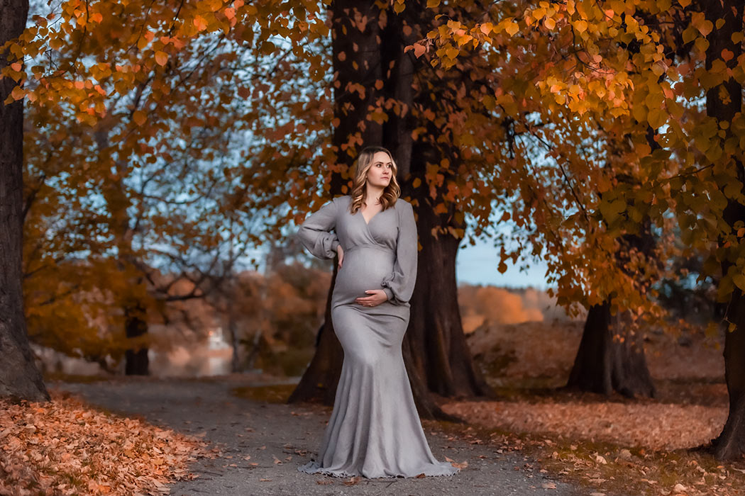 Gravidfotografering i slutet av oktober under stora träd med orange löv i Hagaparken. Den gravida kvinnan har på sig en grå mysig gravidklänning och står på en grusgång som omges av nedfallna orange löv som täcker gräsmattorna.