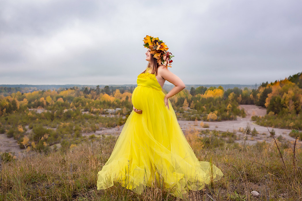 Höstig gravidfotografering i oktober på en höjd ovanför gula och bruna träd. Kvinnan har på sig en gul tyllklänning och på huvudet en krona av höstlöv.