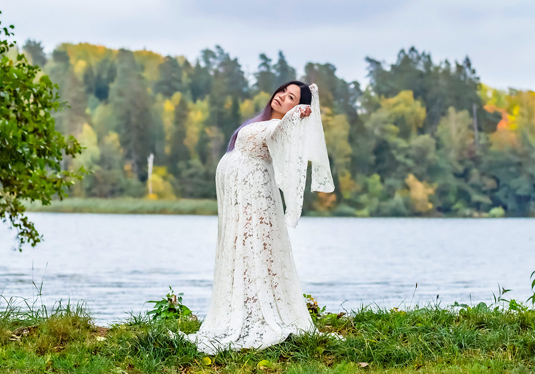Gravidfotografering på hösten vid en sjö med höstfärgade träd runt. Kvinnan har en vit spetsklänning och tittar in i kameran.