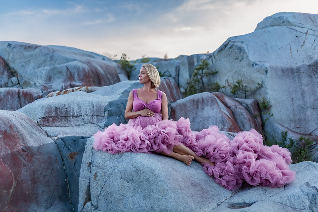 Gravidfotografering i ett stenbrott. Kvinnan har på sig en rosa tyllklänning och sitter bland de stora stenarna.