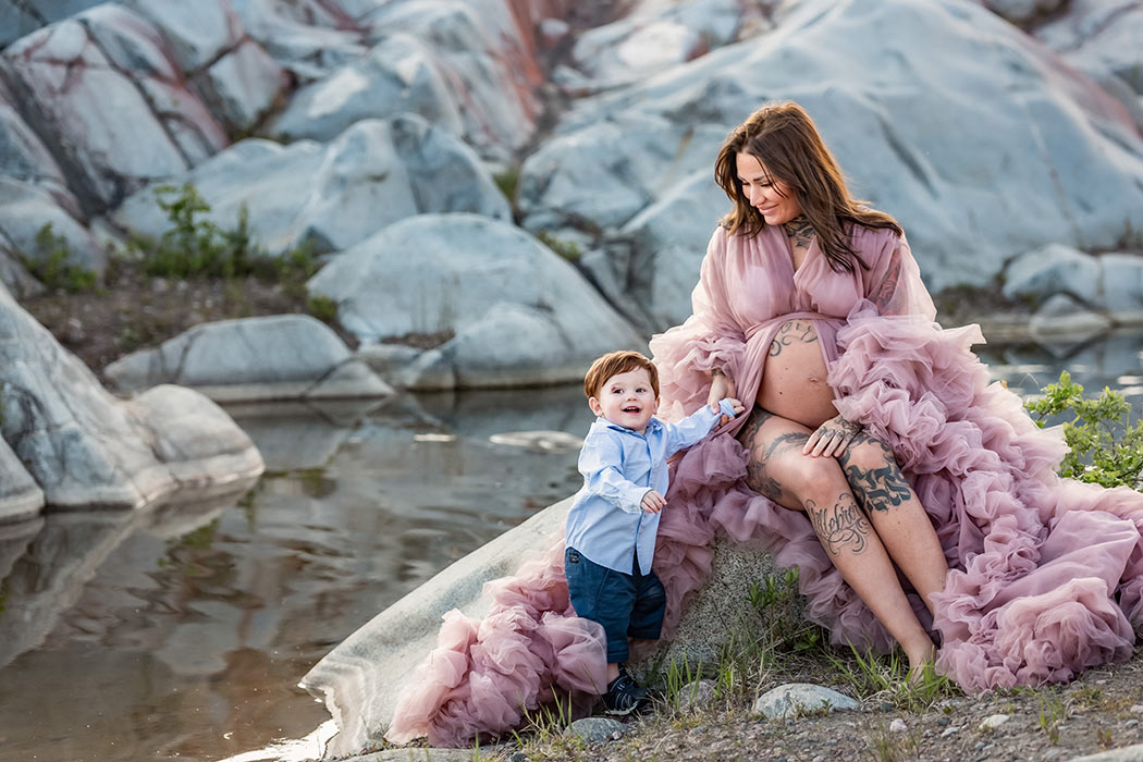 En gravidkvinna sitter på en stor sten och håller hand med sin ettåriga son.