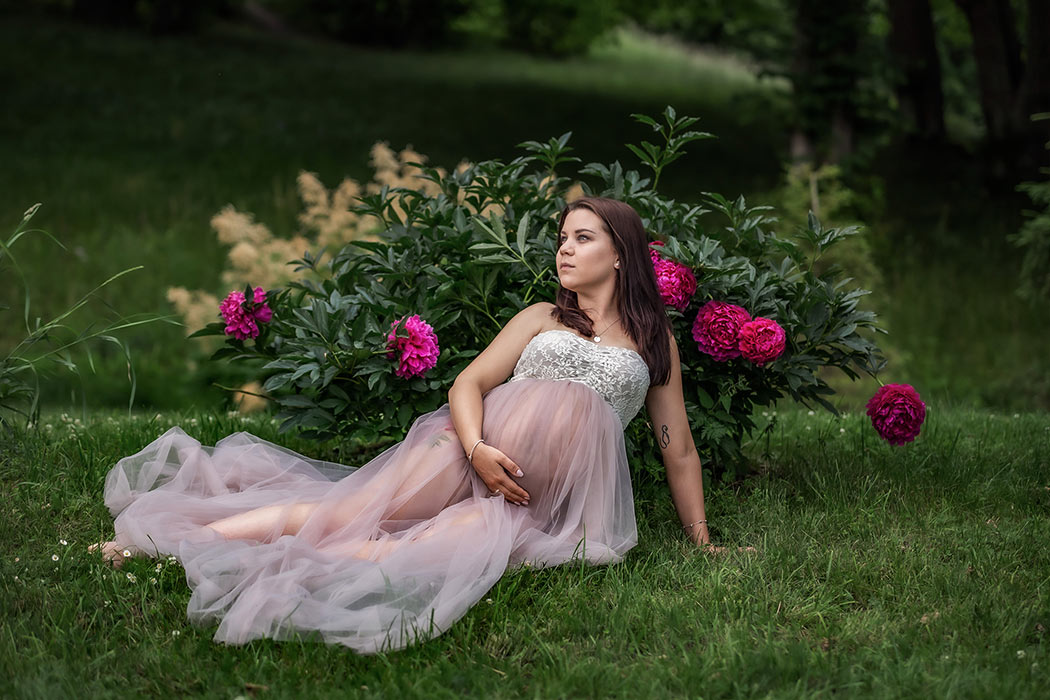 Gravidfotografering på sommaren. Den gravida kvinnan sitter på gräset framför en stor pionbuske med djuprosa blommor.