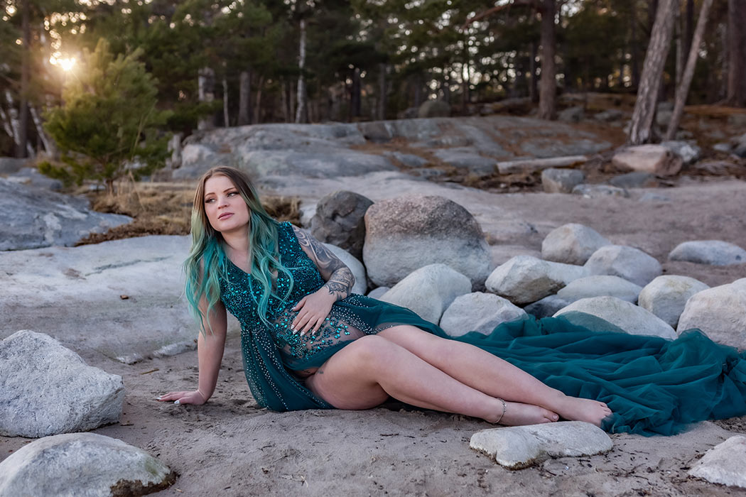 Gravidfotografering på en strand med stora stenar. Den gravida kvinnan har på sig en grön glittrande klänning och sitter på sanden. I bakgrunden syns en tallskog där solen skiner in mellan grenarna.