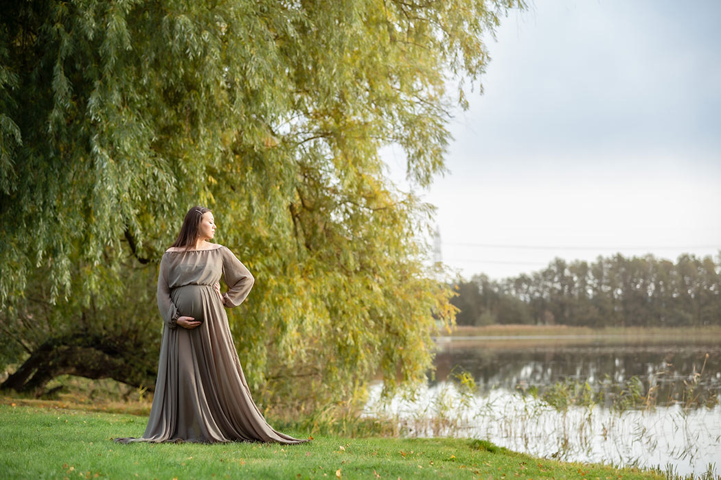 Gravidfotografering i slutet av oktober vid en sjö. Den gravida kvinnan står framför ett gigantiskt pilträd och på andra sidan vattnet syns träd och vass.