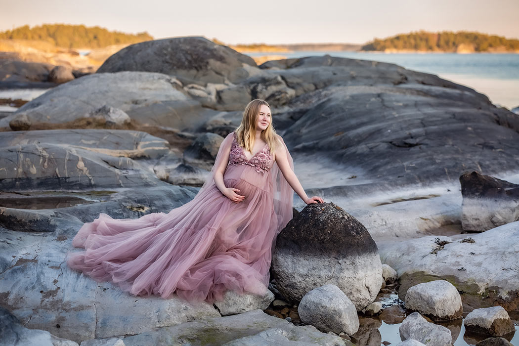 En gravid kvinna sitter bland klippor och stora stenar. I bakgrunden syns havet och kvällssolen som lyser upp öar.