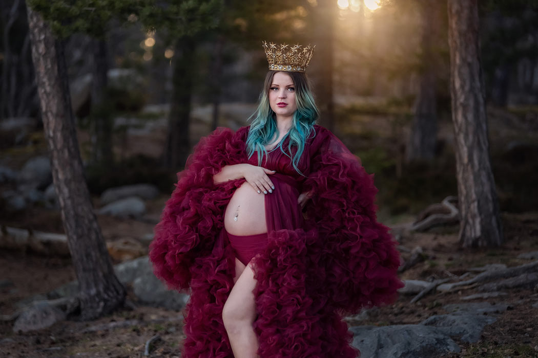 Gravidfotograferingen i en barrskog. Den gravida kvinnan har på sig en röd tyllklänning som är öppen över magen och en krona. Solen strålar in genom grenarna i bakgrunden.