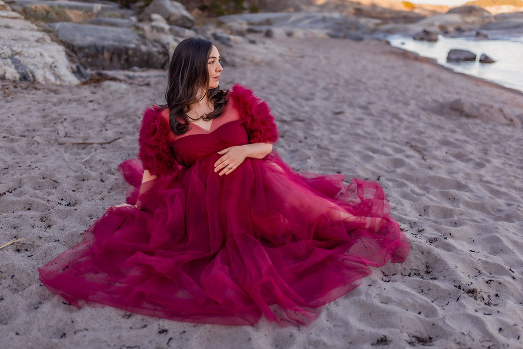 En gravid kvinna i röd klänning sitter på en sandstrand och tittar ut över havet.