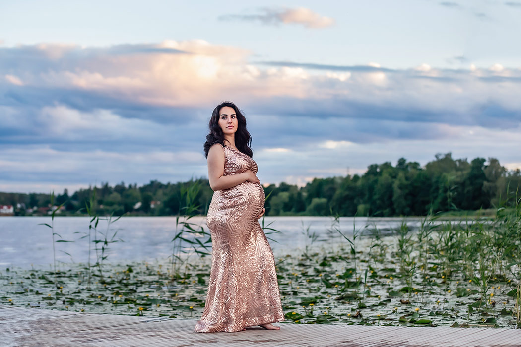 En gravid kvinna i en fantastisk glittrande gravidklänning står på en brygga vid en sjö. I bakgrunden syns näckrosblad och vackra moln i solnedgången.