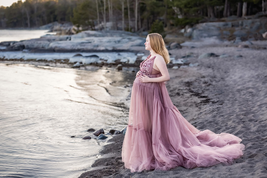 Gravidfotografering med en gravid kvinna i en rosa tyllklänning med blomdetaljer. Kvinnan står på stranden och tittar ut över vattnet.