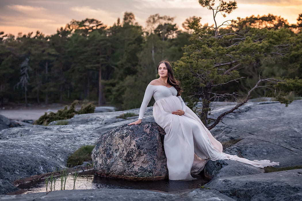Gravid kvinna sitter på en stor sten som ligger i en vattenpöl på klipporna. I bakgrunden syns en skog och en vacker solnedgång.