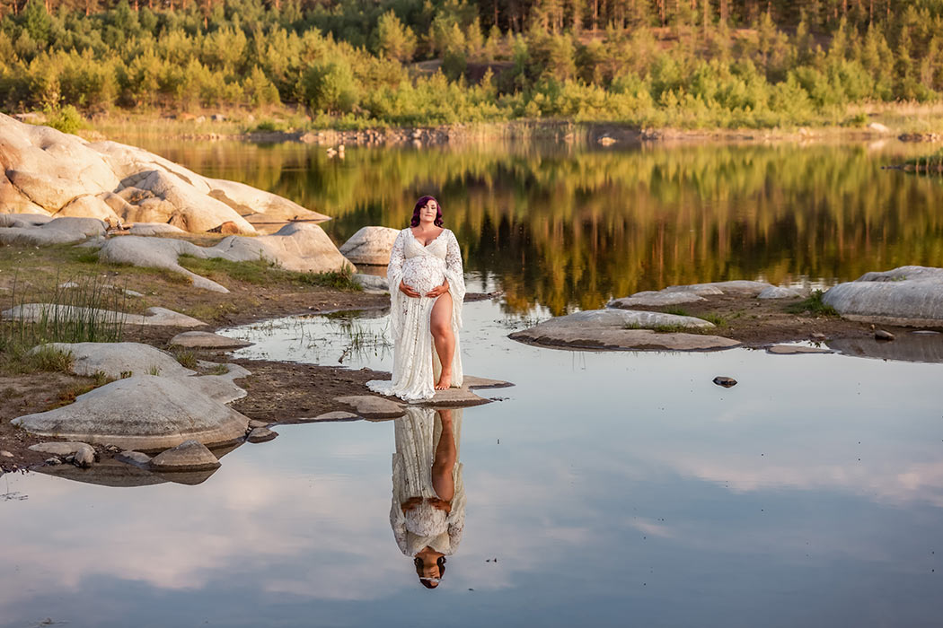 Magiskt vacker gravidfotografering i ett spegelblankt vattenlandskap omgivet av klippor och skog. Den gravida kvinnan har en vit spetsklänning och står utmed vattnet.