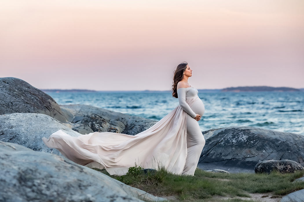 En gravidkvinna i en beige klänning med långt släp står på klipporna vid havet. Himlen är rosafärgad av solnedgången.