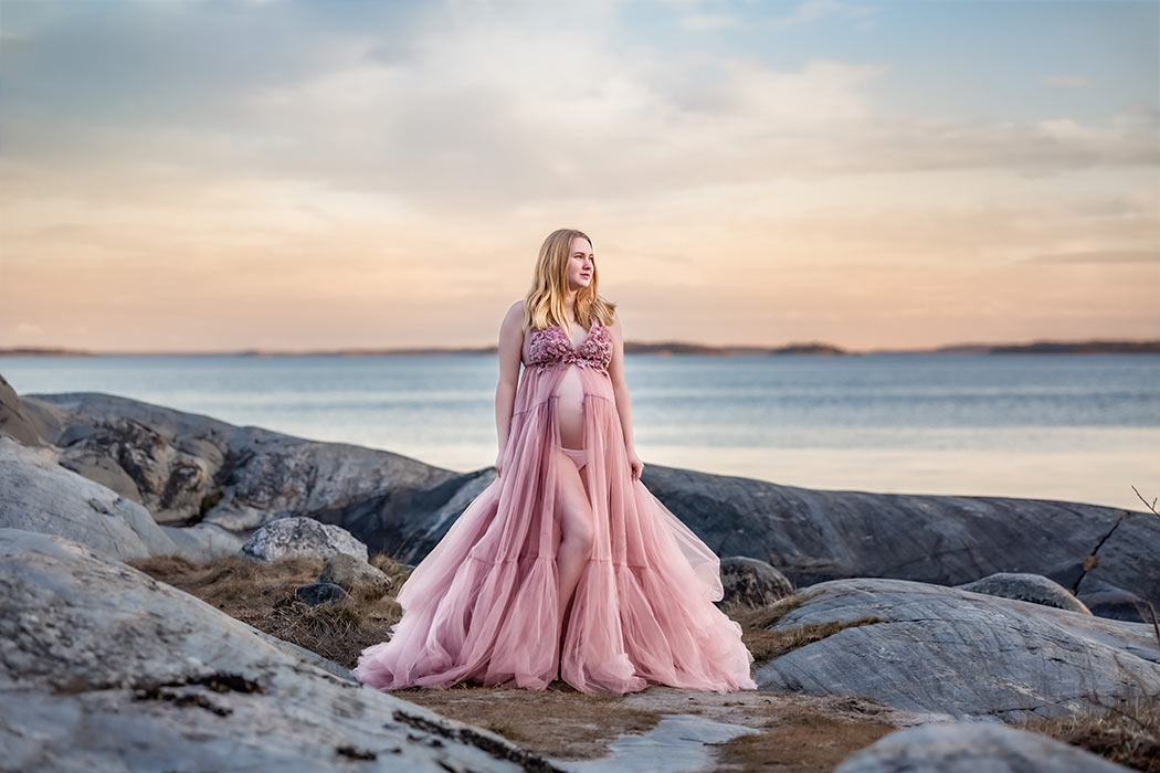 En gravid kvinna står på klippor med havet i bakgrunden. På sig har hon en rosa tyllklänning som är öppen för att visa magen.