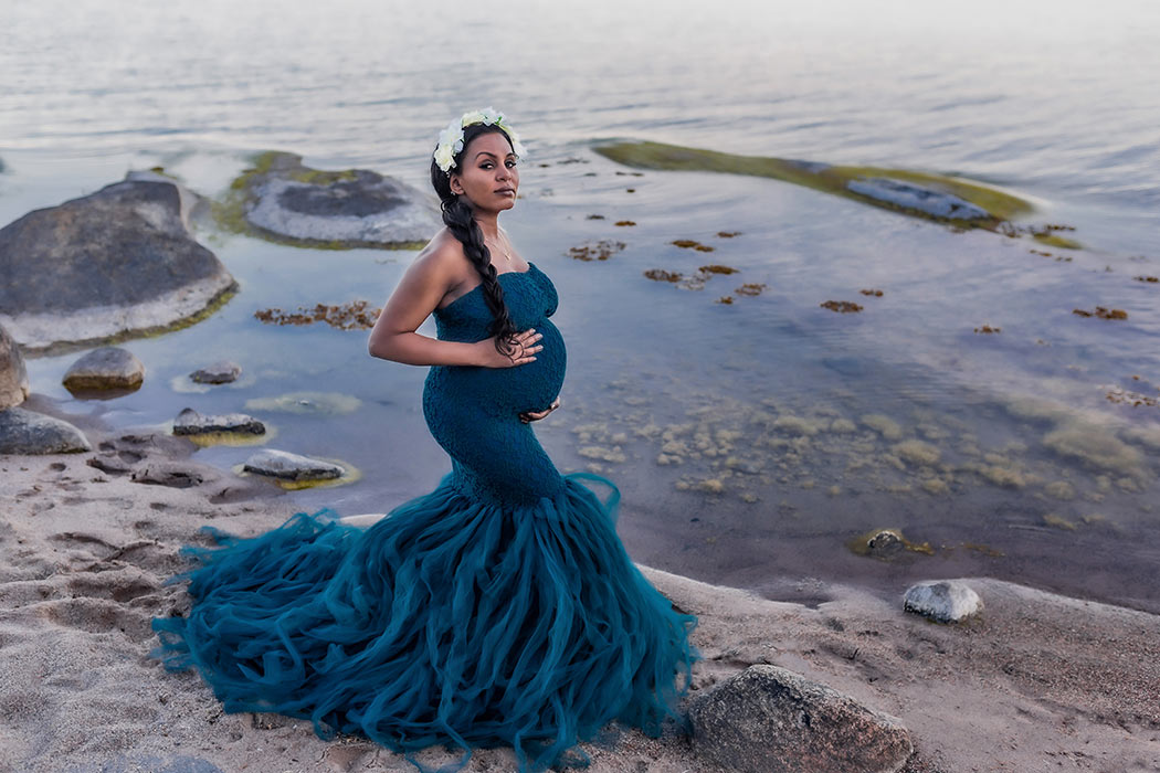 En vacker gravid kvinna har på sig en blå klänning av spets och tyll. Hon står på stranden i bakgrunden syns vatten och stenar med sjögräs.