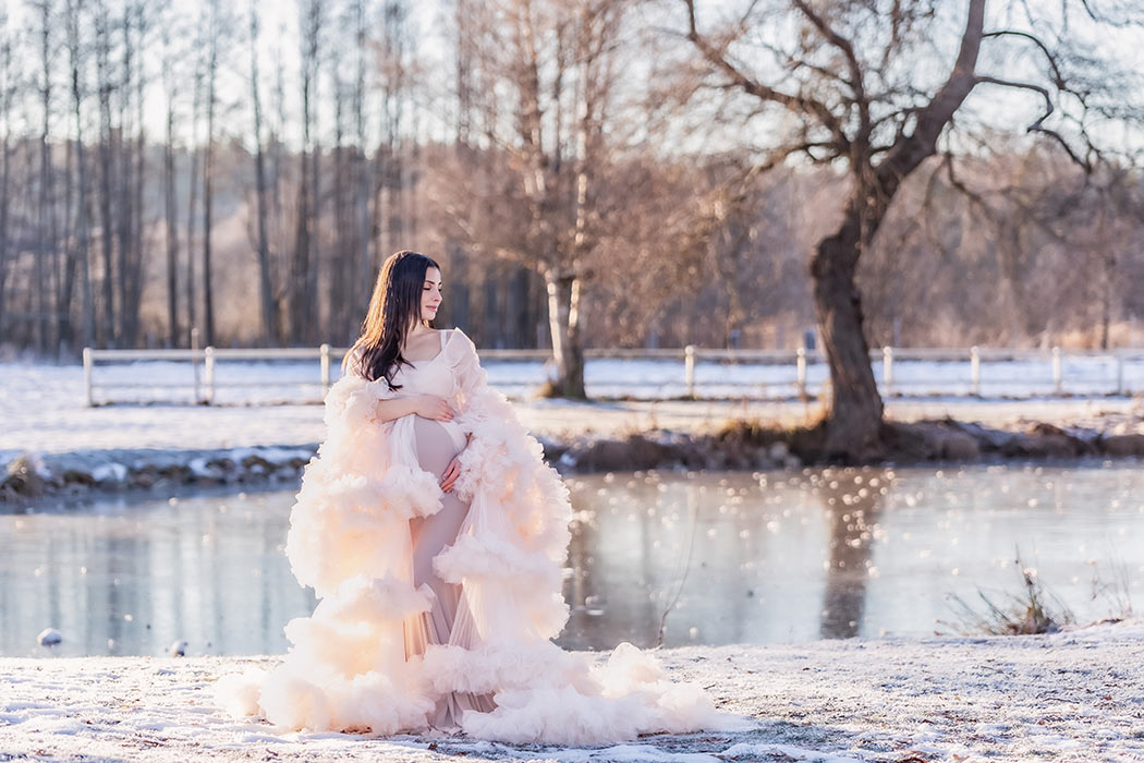 Gravidfotografering en kall vinterdag vid en isig sjö. Den gravida kvinnan har på sig en vacker ljusbeige tyllklänning.