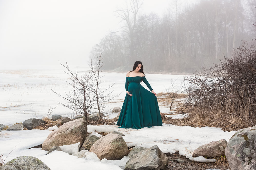 En gravid kvinna i grön klänning står i ett vinterlandskap där dimman ligger tät.