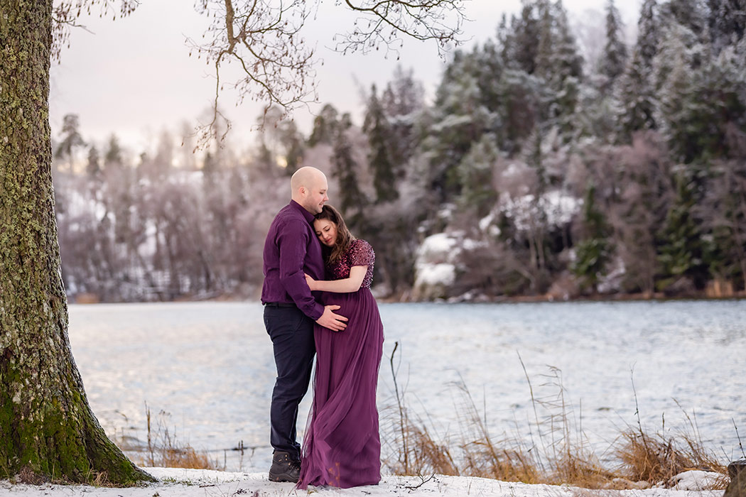 Gravidfotografering i ett snölandskap vid en glittrande sjö. Den gravida kvinnan kramar om den blivande pappan under ett träd. Båda har på sig lila kläder.