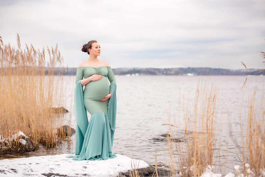 Gravidfotografering på vintern. Den gravida kvinnan har en vacker grön gravidklänning och står på en snötäckt sten bland vass vid havet.