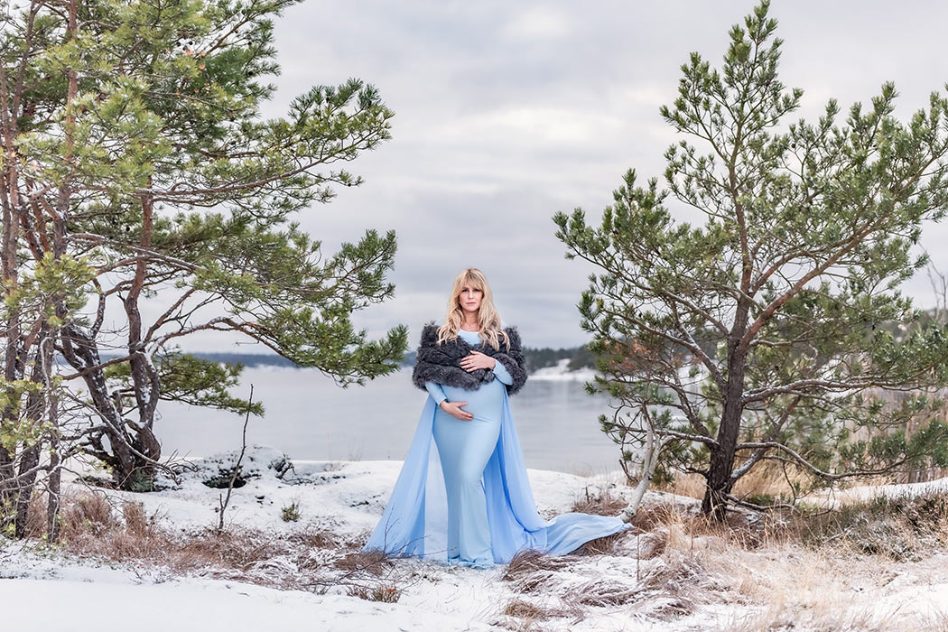 Gravidfotografering vid havet en snövit vinterdag. Den gravida kvinnan har en blå gravidklänning och står mellan två barrträd. I bakgrunden syns det isiga havet.