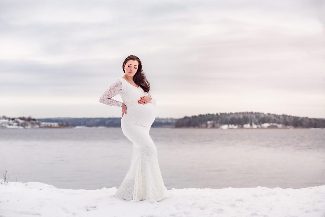 Fotografering en vinterdag med tjock snö vid havet. Vid vattenkanten står en gravid kvinna i vit spetsklänning.