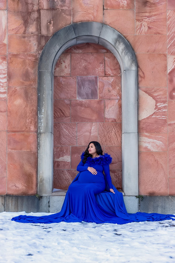 Gravidfotografering där den gravida kvinnan har blå klänning och sitter vid en rosa vägg.