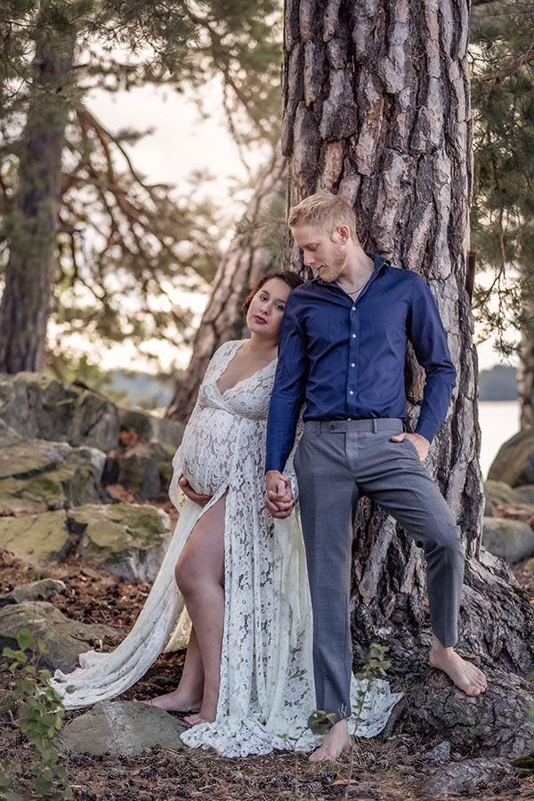 Gravidfotografering med ett par som står lutade mot en stor tallstam i ett stenigt landskap.