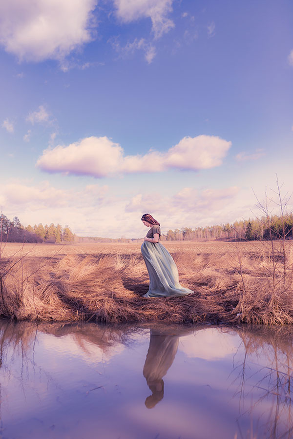 En gravid kvinna står bland grånande höga gräs vid kanten av ett stilla vatten. Hennes reflektion är tydlig i vattnet och himmlen är blå med små vita moln.
